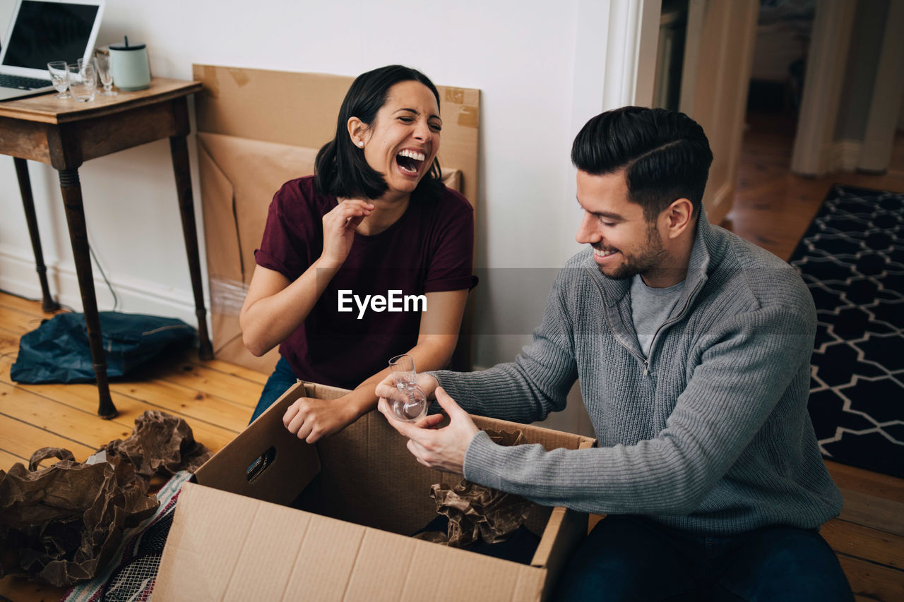 Laughing couple unpacking cardboard box while sitting on floor at home