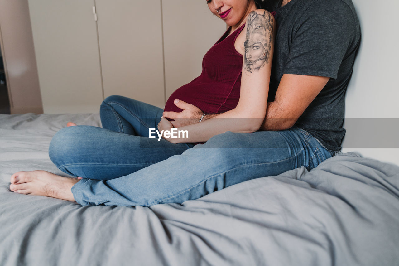 Midsection of pregnant woman sitting with male partner on bed at home