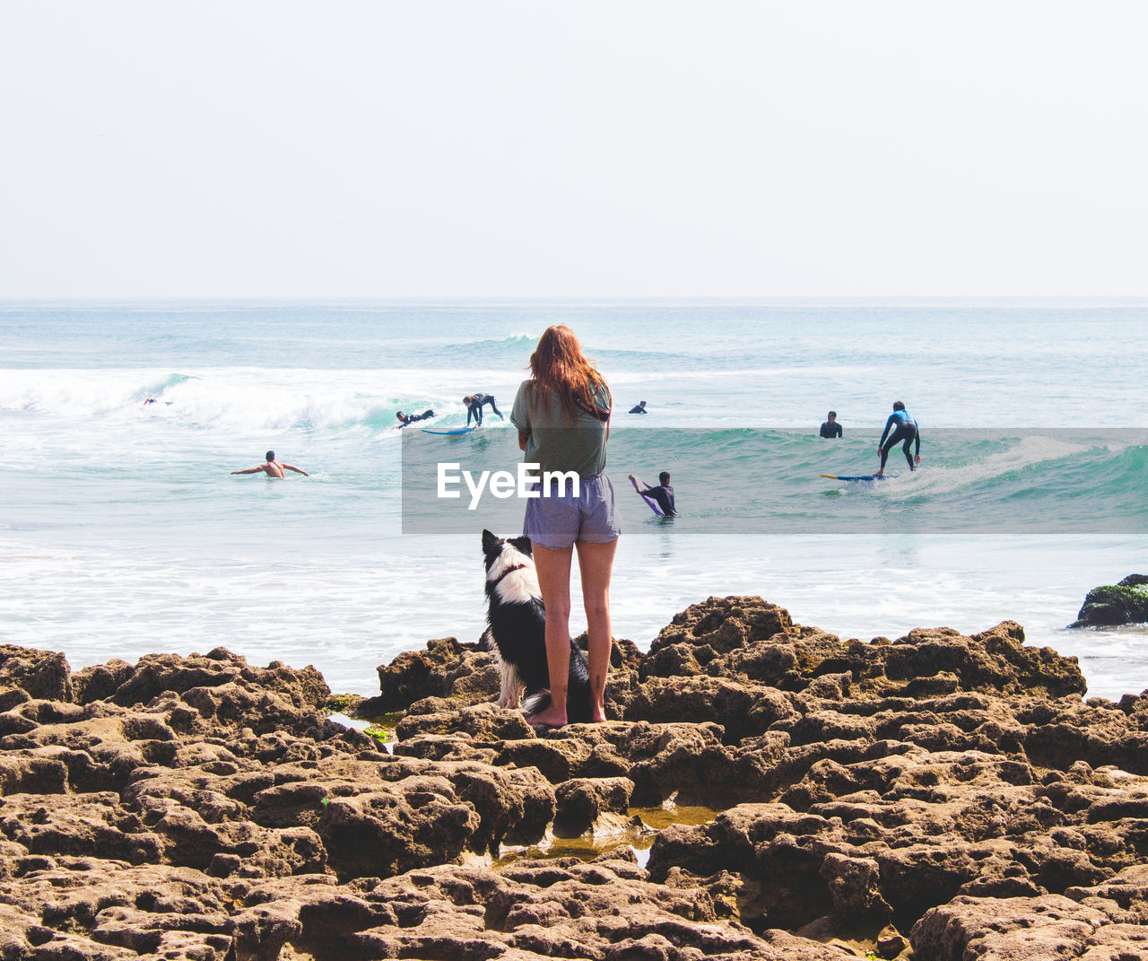 REAR VIEW OF PEOPLE ON ROCK AT BEACH AGAINST SKY