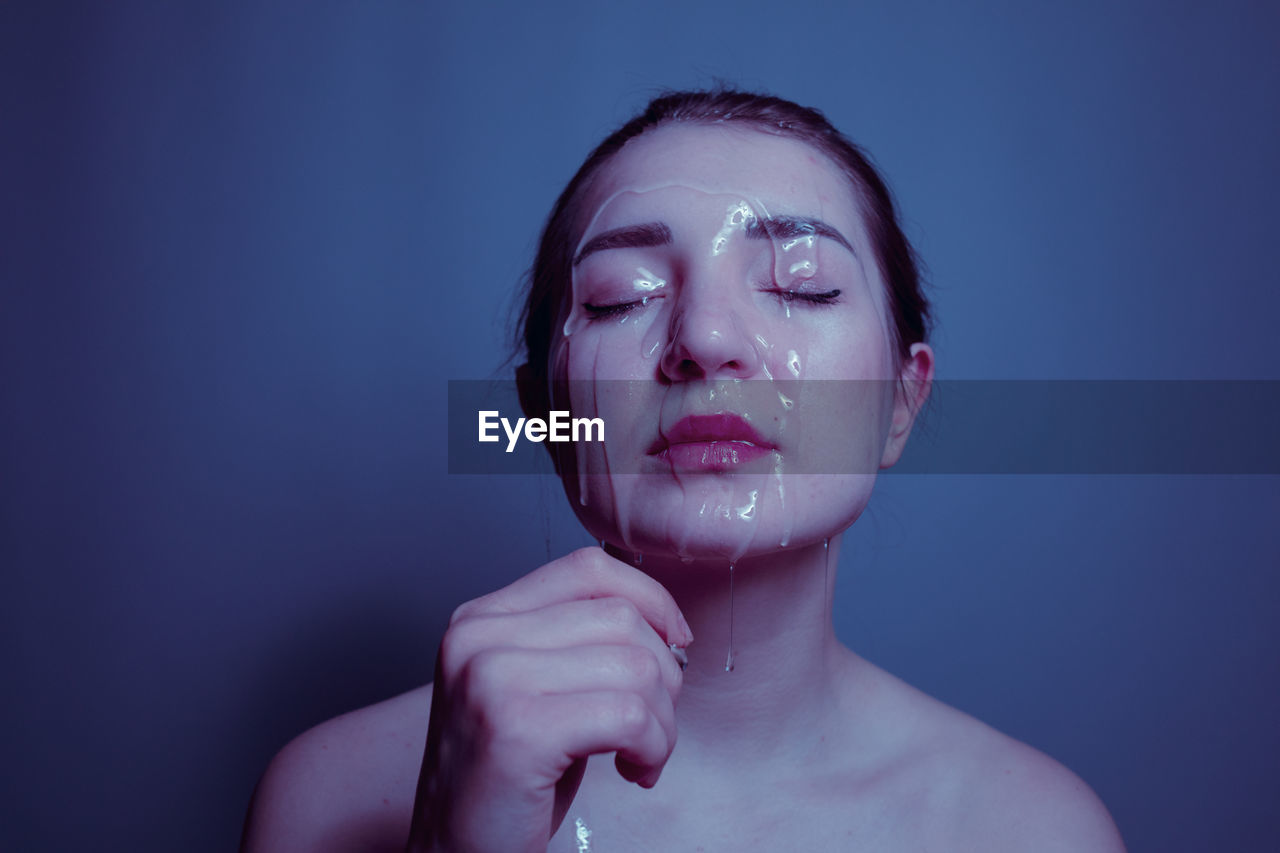 Close-up of shirtless young woman with beauty product on face against blue background