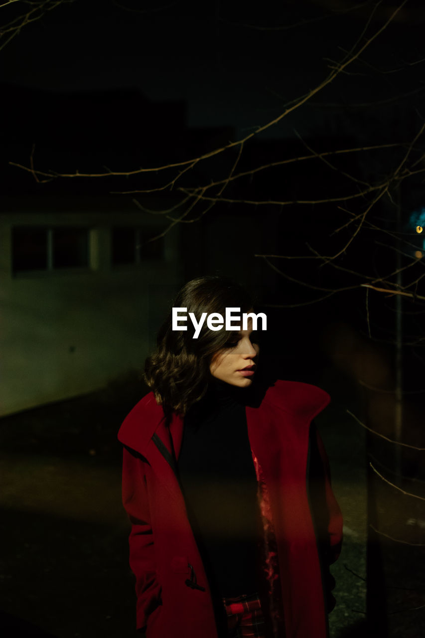 YOUNG WOMAN LOOKING AWAY WHILE STANDING ON ILLUMINATED TREE