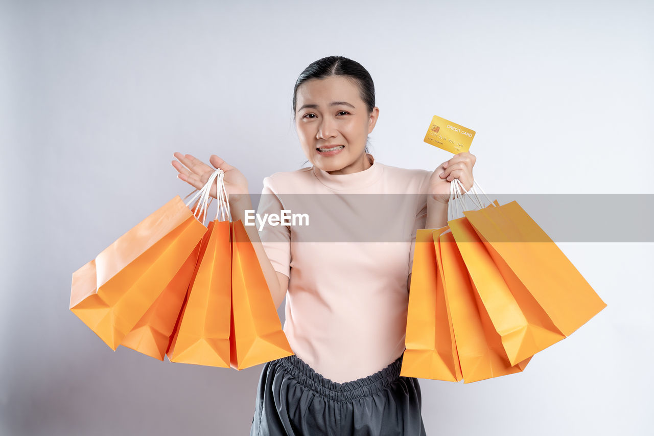 shopping bag, shopping, consumerism, bag, spending money, happiness, orange, retail, smiling, yellow, holding, paper bag, one person, tote bag, cheerful, customer, studio shot, emotion, women, carrying, buying, adult, handbag, fashion accessory, celebration, clothing, paper, portrait, casual clothing, young adult, gift, sale, holiday, positive emotion, front view, lifestyles, indoors, standing, looking at camera, fun, female, copy space, business, enjoyment, smile, excitement