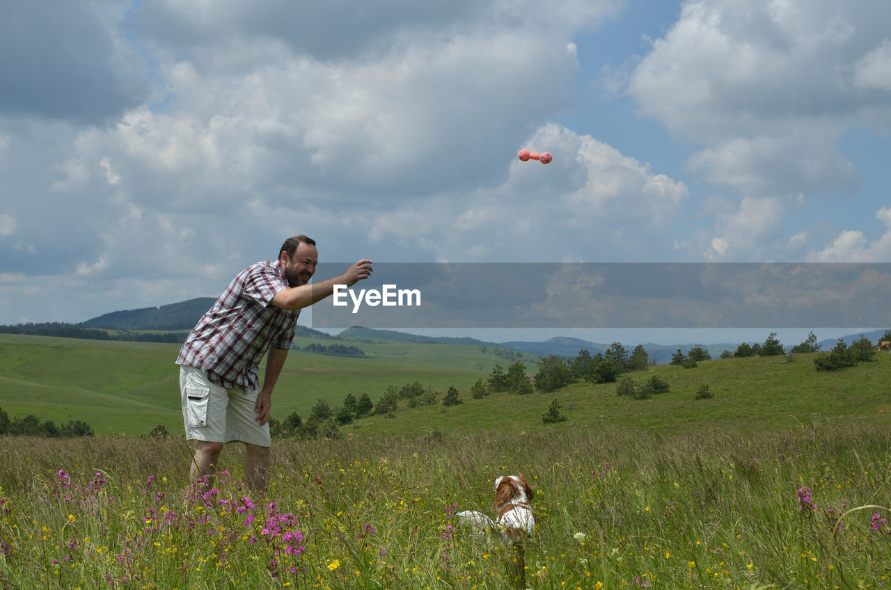 Man is playing with his dog on a meadow, surrounded with pink flowers and with hills in background