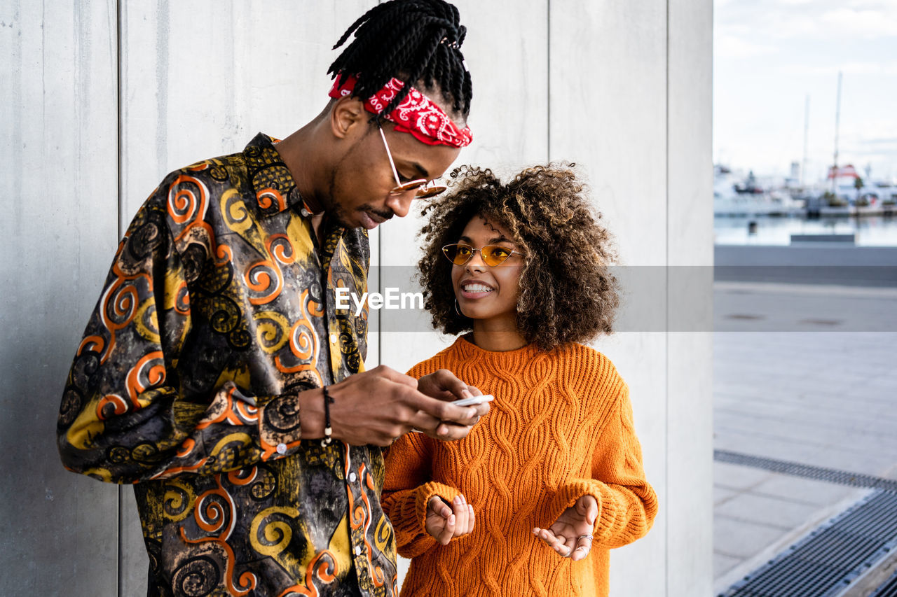Cheerful black woman with curly hair standing with cool ethnic guy in sunglasses and smiling while using mobile phone against gray wall in city