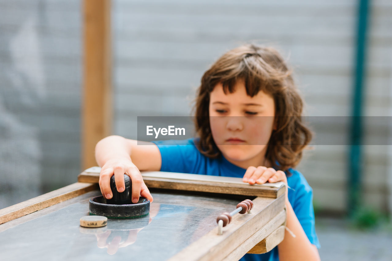 Attentive little girl with brown hair in blue t shirt playing air hockey in entertainment center in daylight