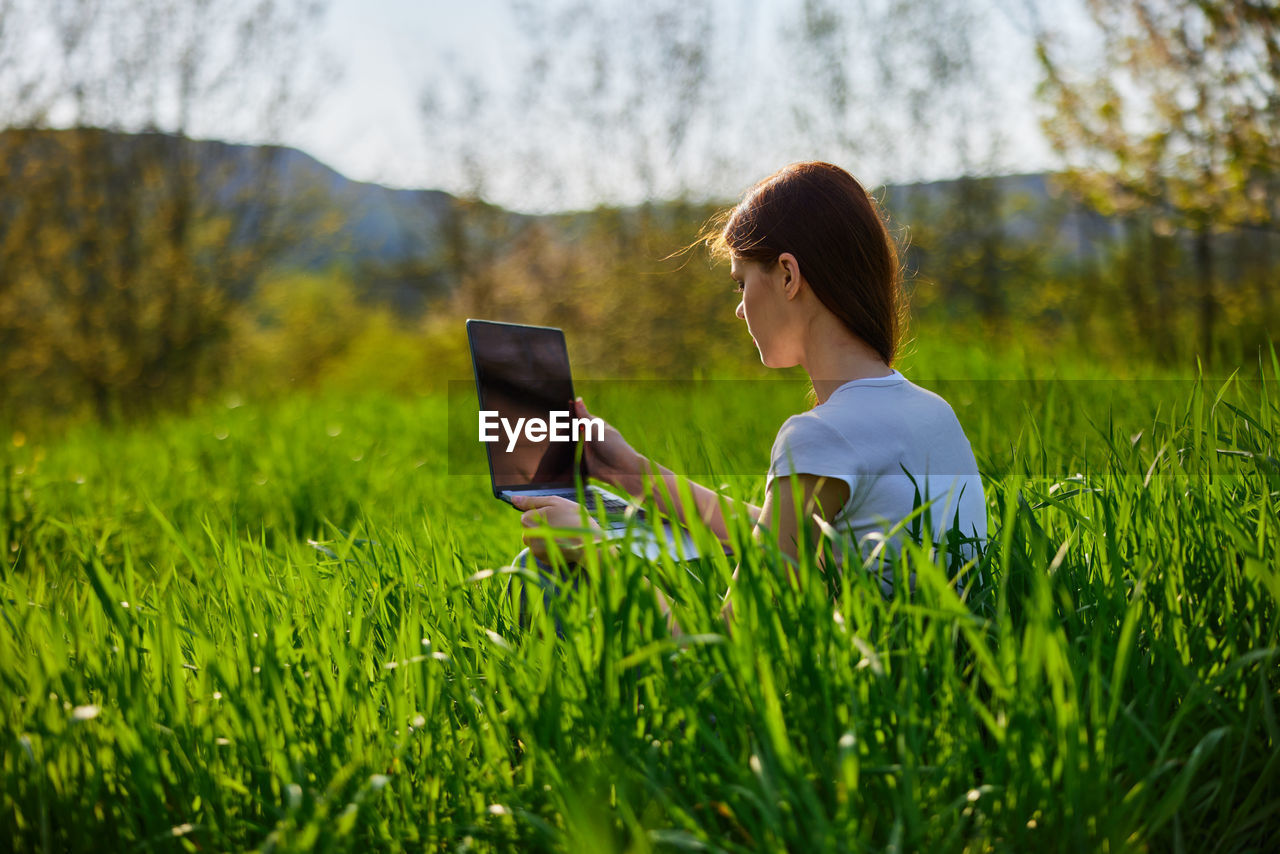 rear view of woman using digital tablet while sitting on grassy field