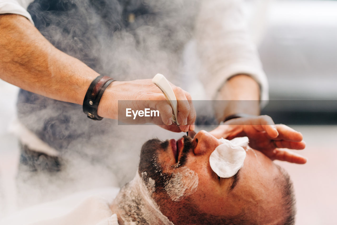 Crop anonymous beauty master shaving beard of client with straight razor during steam vapor treatment in hairdressing salon