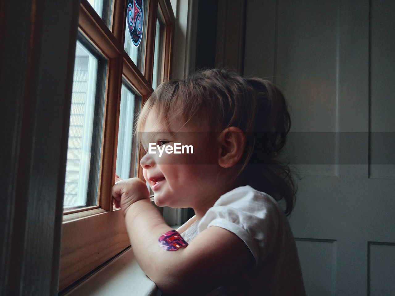 Side view of cute smiling baby girl looking through window at home