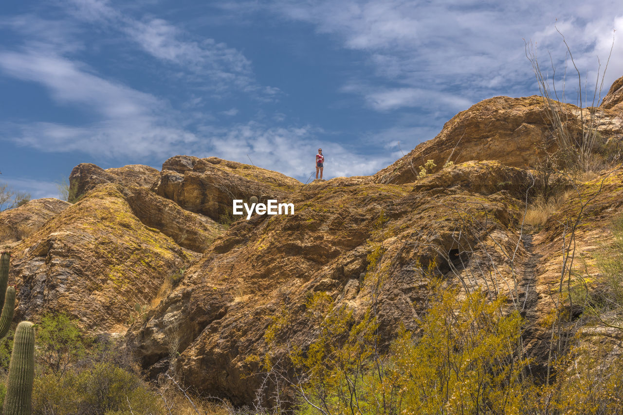 LOW ANGLE VIEW OF PERSON STANDING ON ROCKS AGAINST SKY