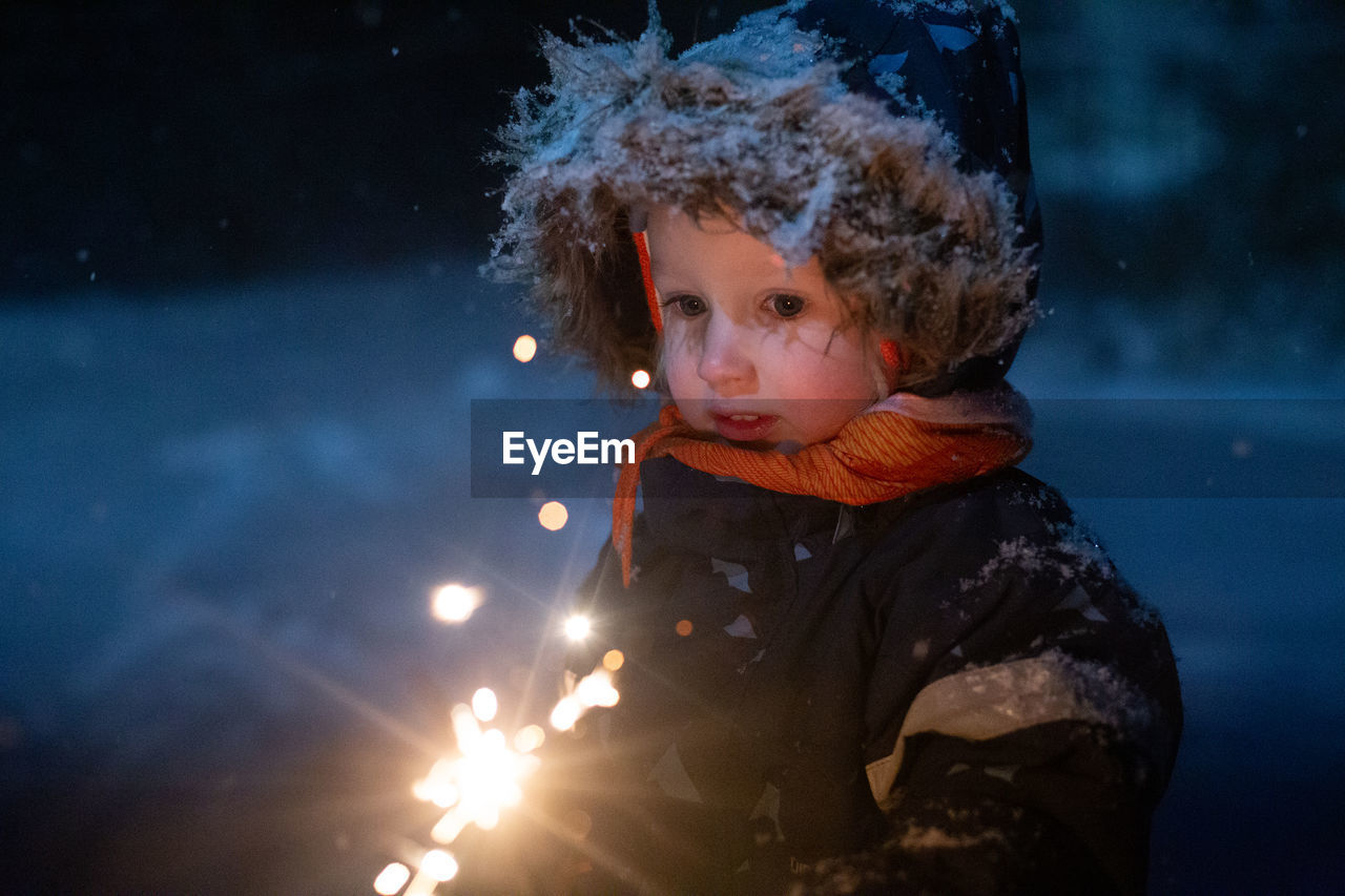 Toddler girl in winter clothes walking outside and holding sparkler in her hand. dark and snowy