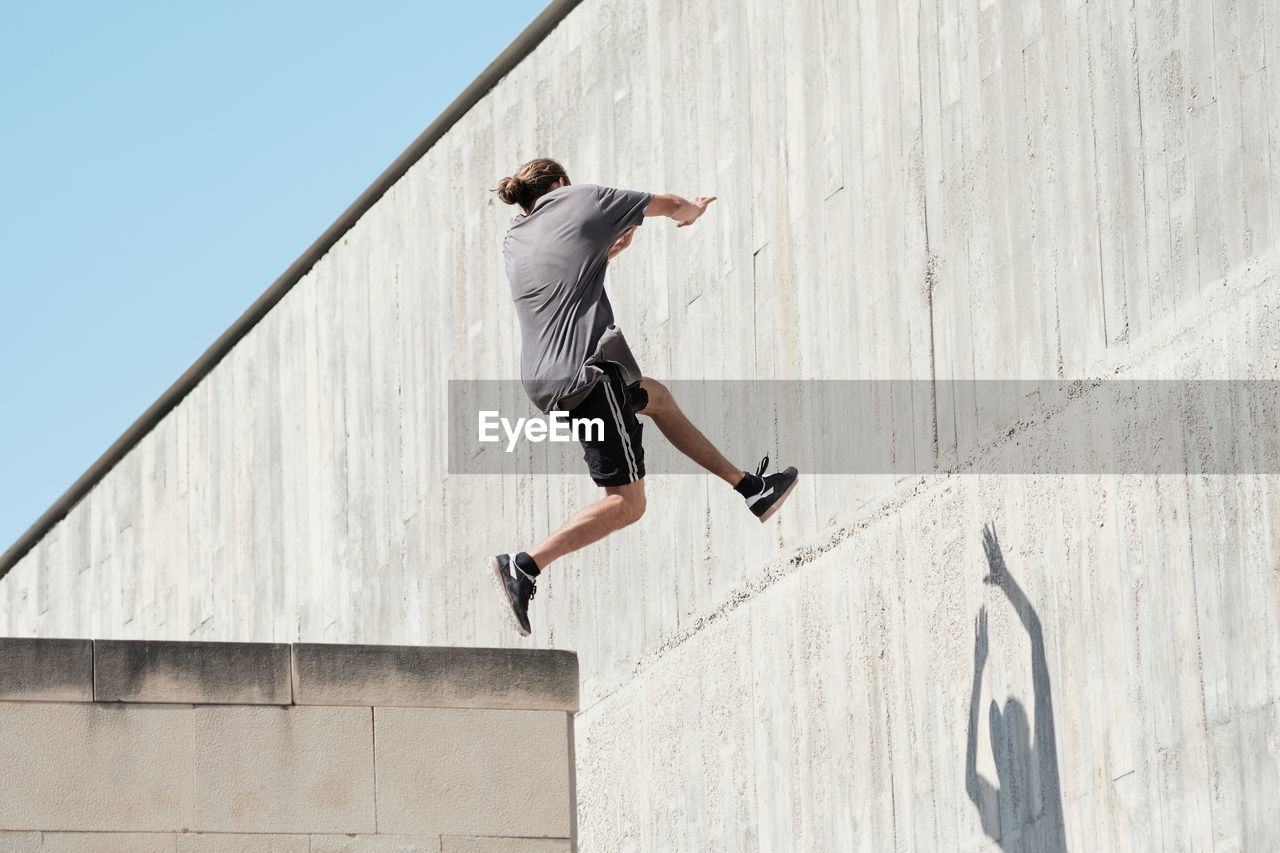 Side view of courageous young male jumping on concrete wall of building while performing dangerous stunt and doing parkour in city