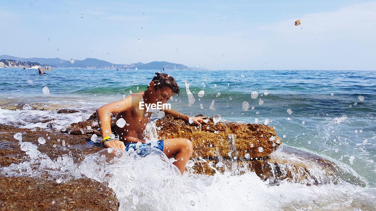 Happy shirtless boy sitting on rock with splashing wave in sea