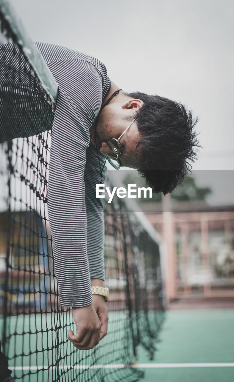 Side view of young man hanging on net against cloudy sky