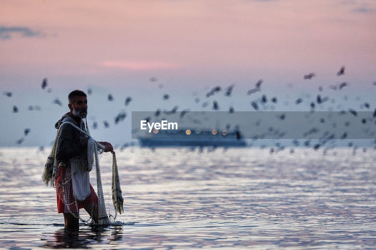 Fisherman standing in sea against sky during sunset