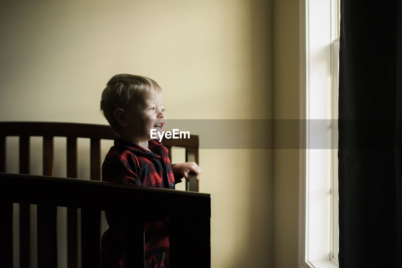 Smiling baby boy looking through window while standing in crib at home