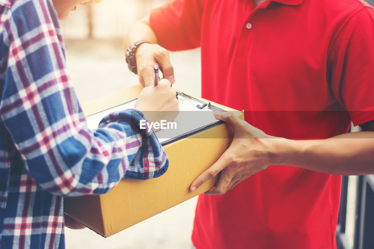 Midsection of female customer signing receipt while receiving package from delivery man outdoors