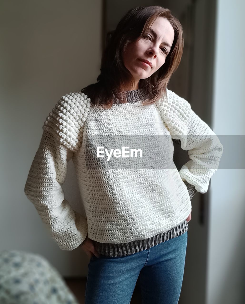 one person, adult, outerwear, sweater, sleeve, women, indoors, clothing, three quarter length, casual clothing, standing, brown hair, portrait, hairstyle, emotion, young adult, long hair, photo shoot, female, jeans, smiling, pattern, fashion, looking at camera, front view, happiness, looking, lifestyles, collar, white, home interior, person, textile, cardigan, cardigan sweater, knitting