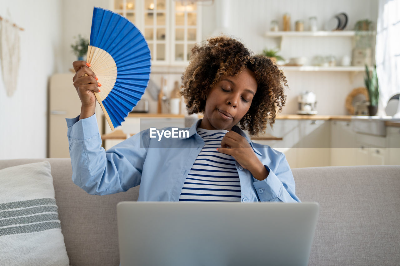 Exhausted overheated african american girl freelancer waving hand fan and working on laptop at home