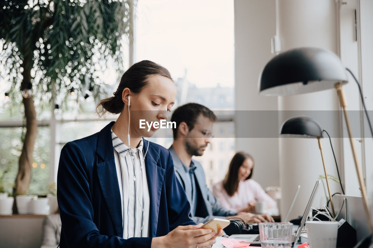 Businesswoman listening music through headphones while sitting with colleagues at desk in office