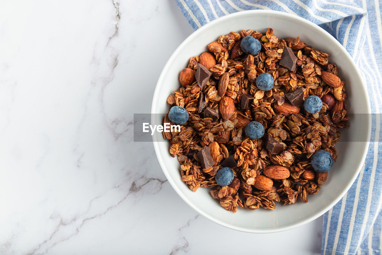Homemade chocolate granola with almonds, hazelnuts and blueberries on white marble background.