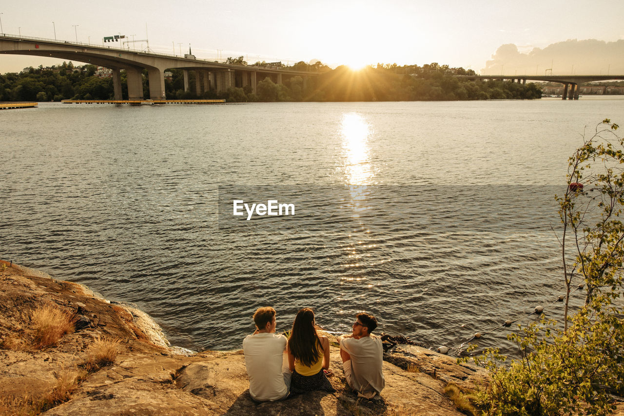 High angle view of friends sitting and talking on rock near sea at sunset