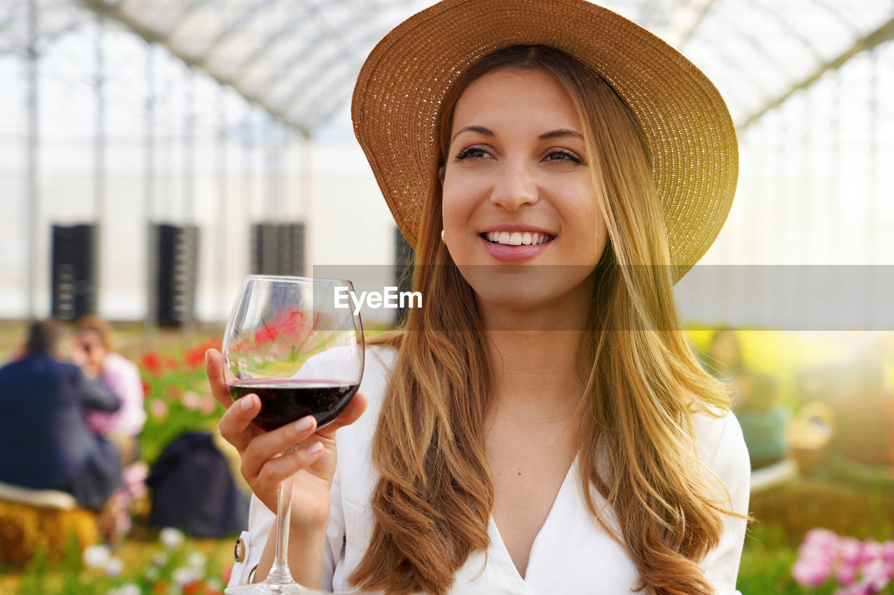 Image of cute young woman enjoying sitting in greenhouse holding glass drinking wine