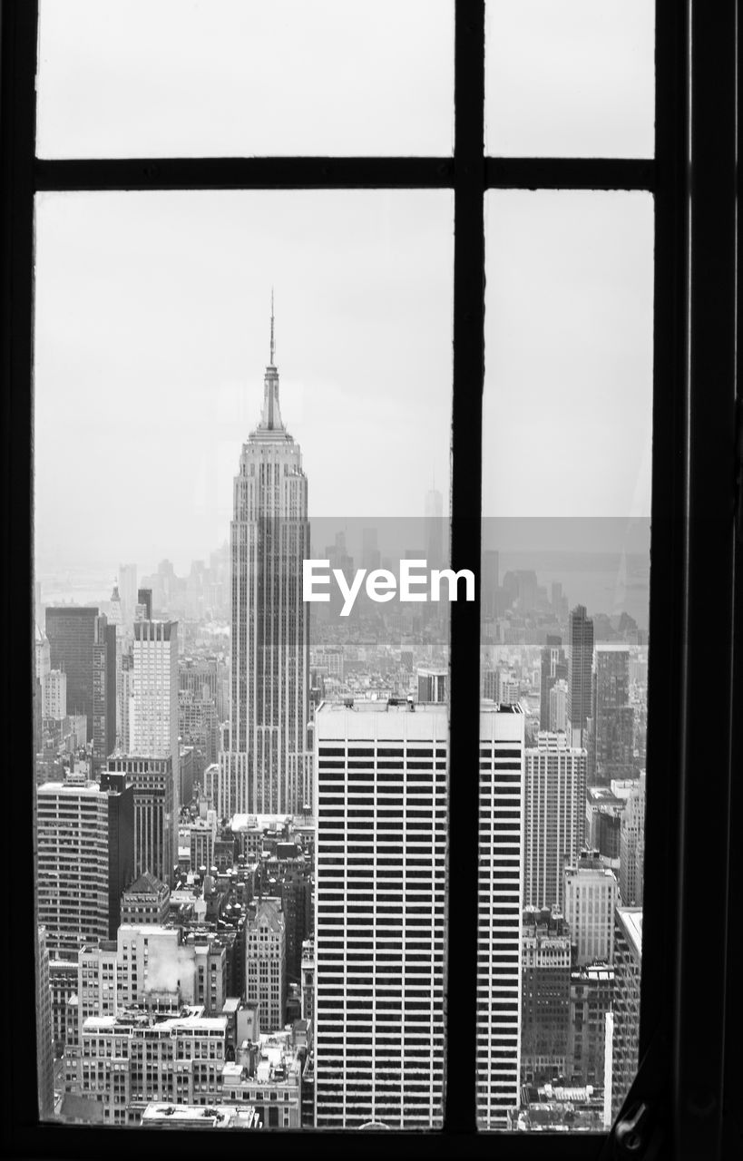 Empire state building in city against sky seen through glass window