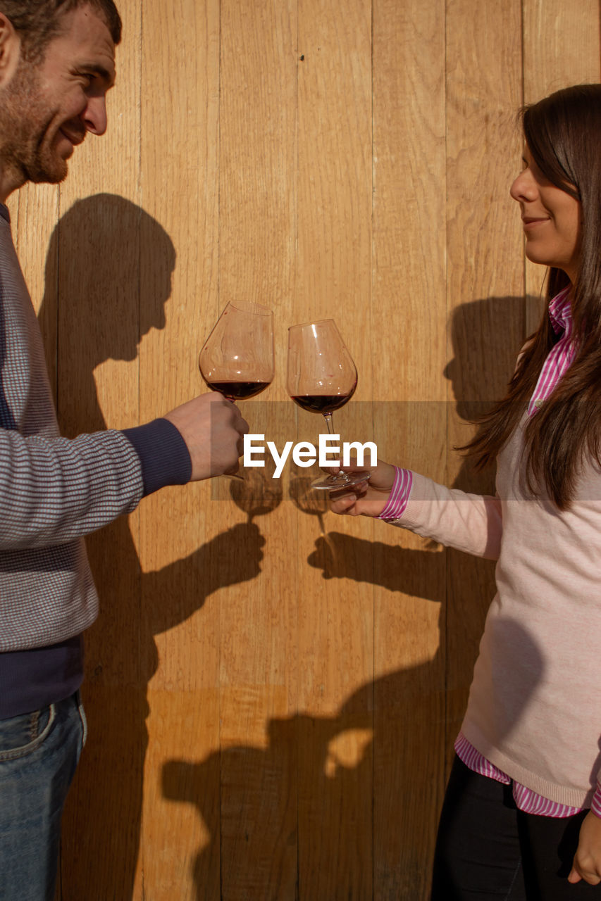 Couple stand with glasses of wine opposite each other on a wooden background with beautiful shadows