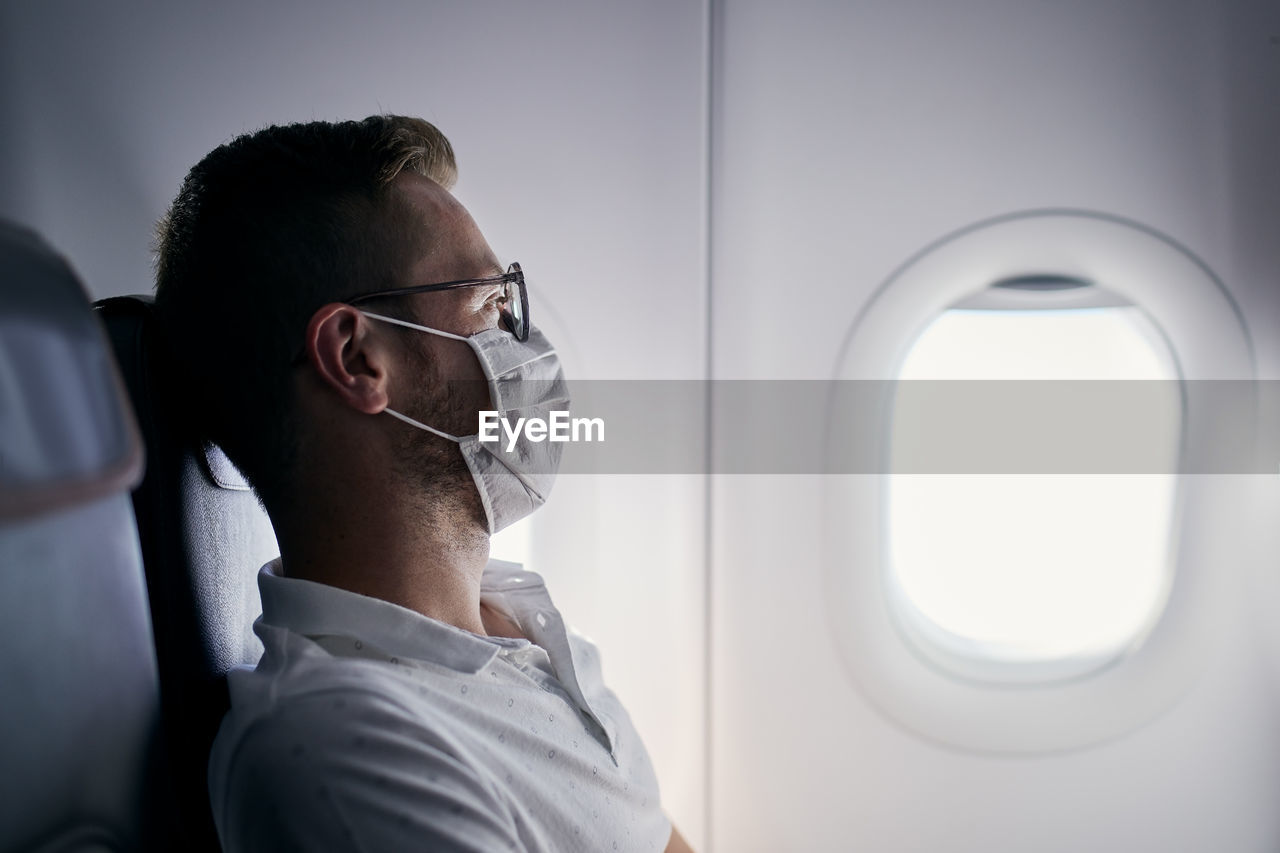 Passenger with protective face mask. young man man looking out window of airplane. 