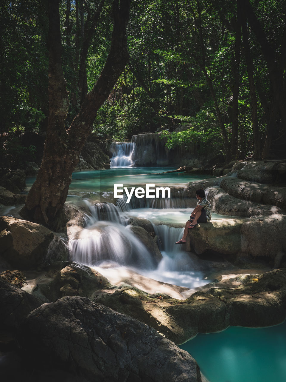 Solo traveler sit on the rock enjoy peaceful scenic waterfall. erawan falls, kanchanaburi, thailand.