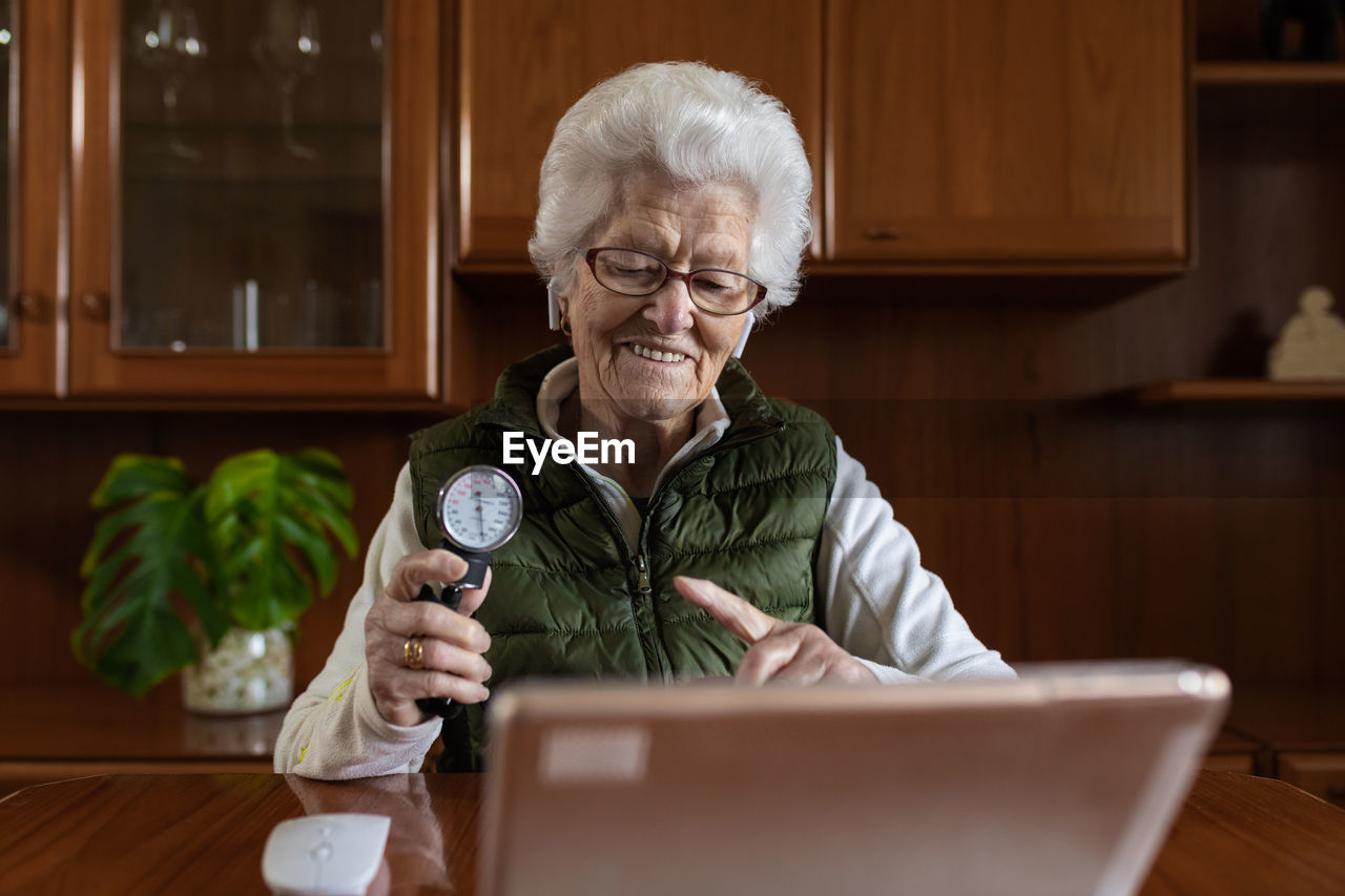 Smiling senior female pensioner pointing at sphygmomanometer while talking on video chat against tablet during online health consultation at home