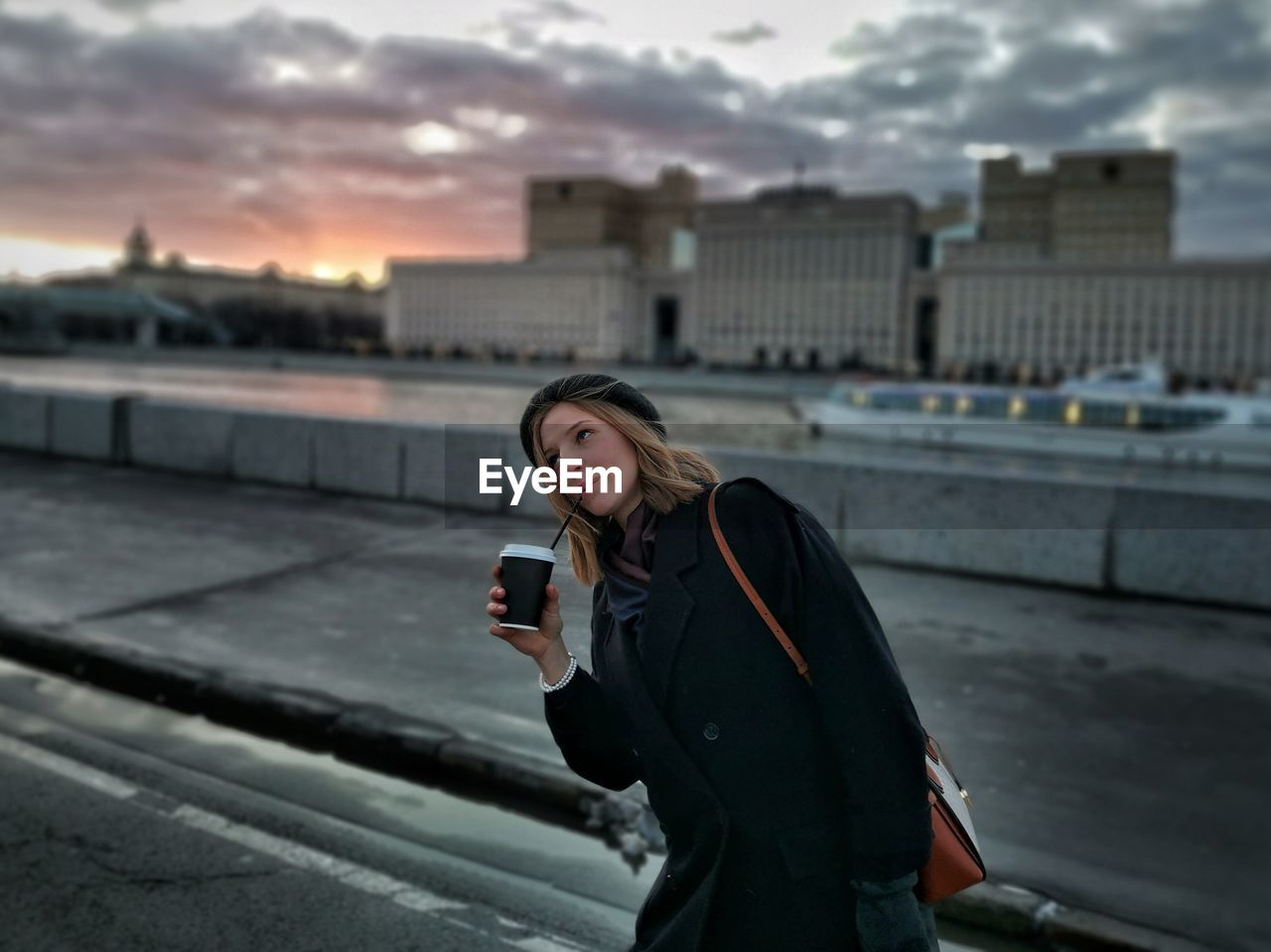 Woman drinking coffee while standing on road in city at sunset