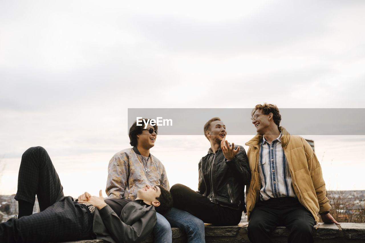 Young male friends spending leisure time while sitting on retaining wall against sky