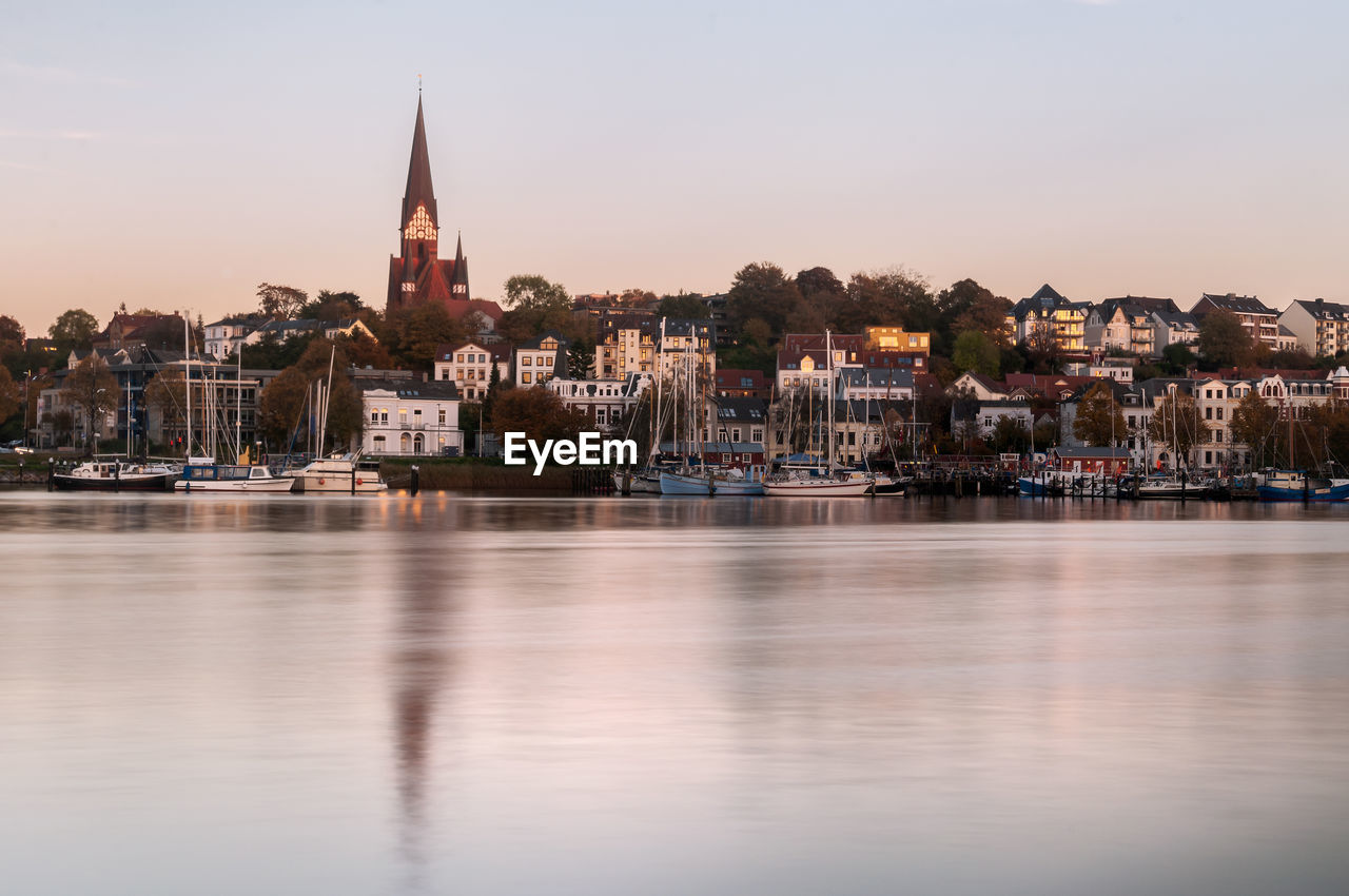 Blurred motion of river by buildings in city against sky during sunset