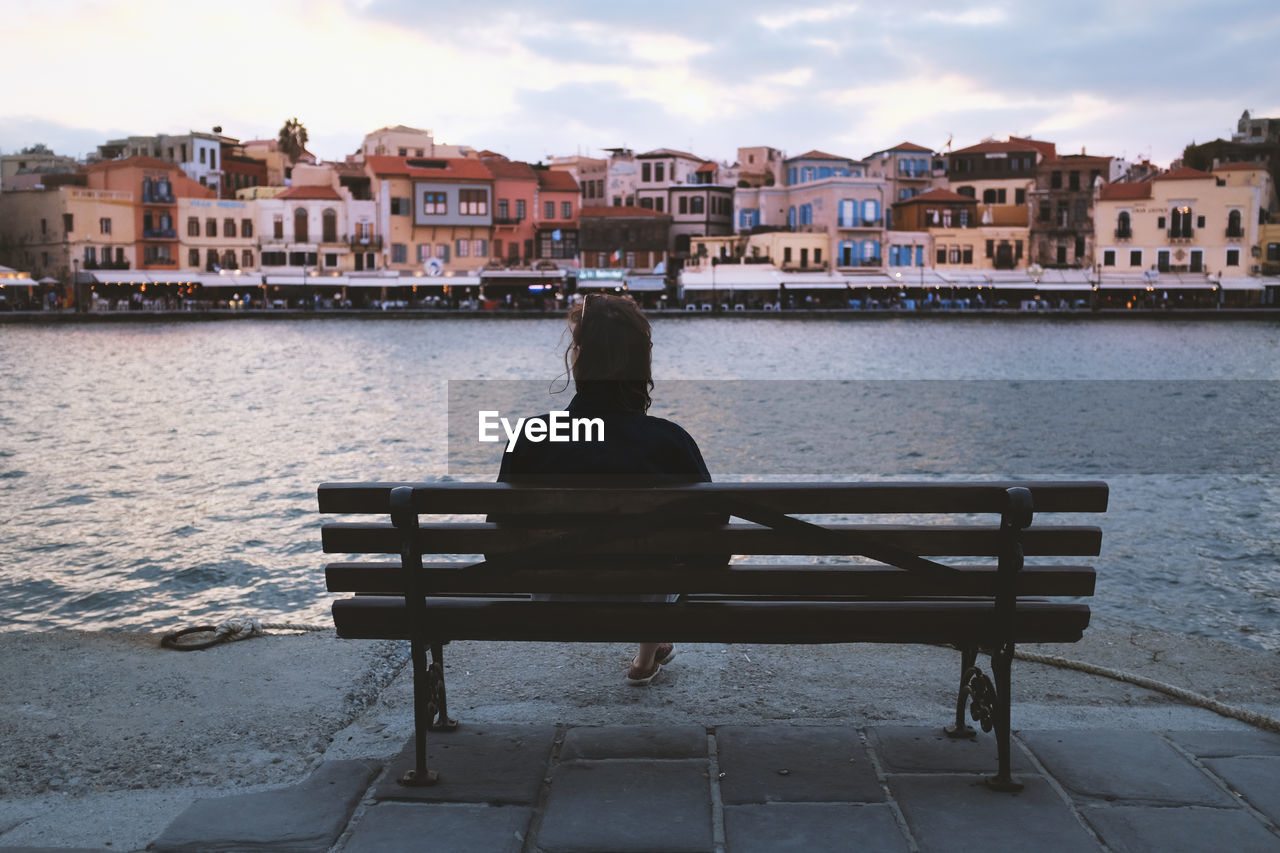REAR VIEW OF MAN SITTING ON BENCH AGAINST BUILDINGS