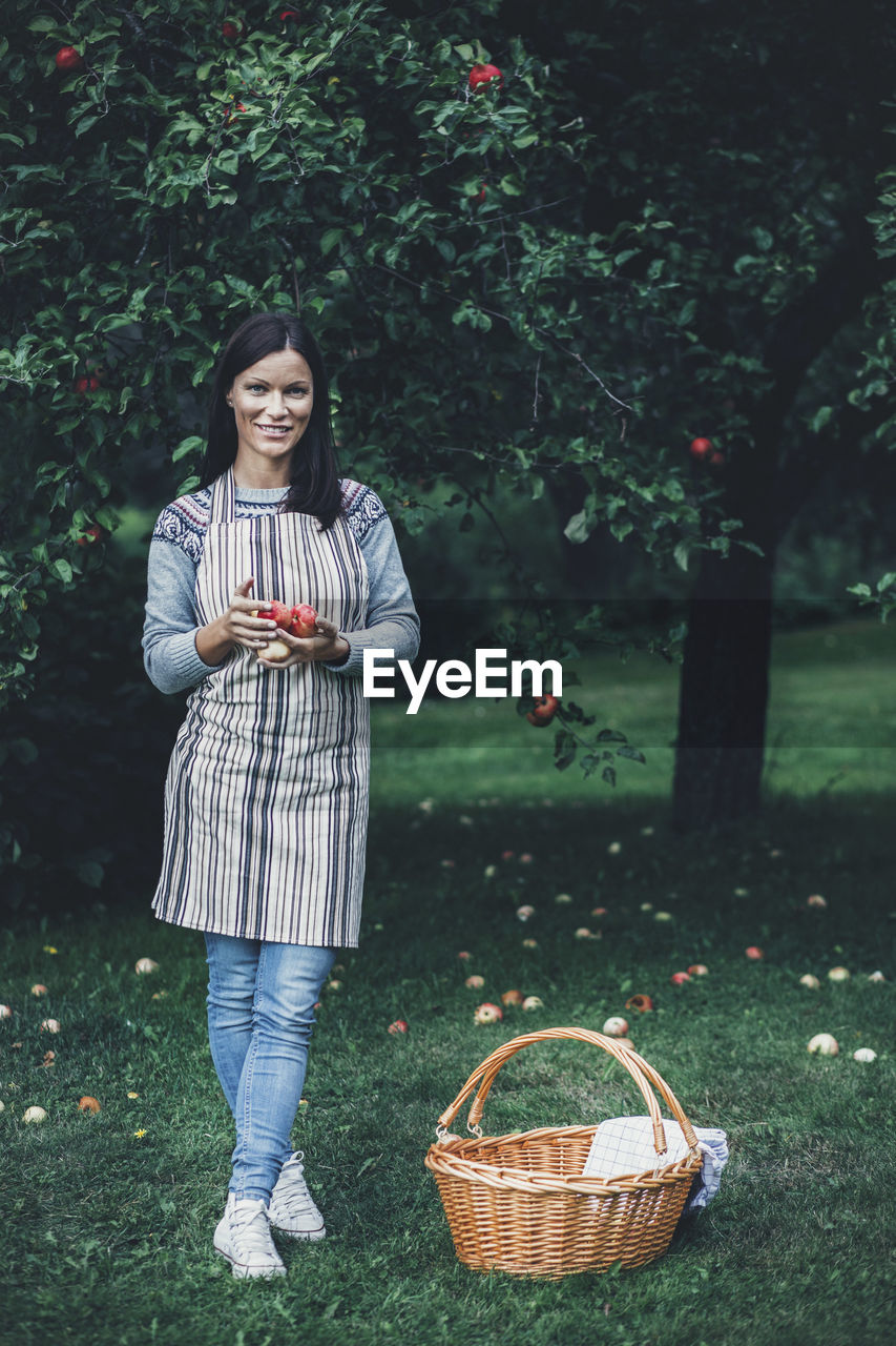 Full length portrait of smiling woman holding fresh apples at orchard