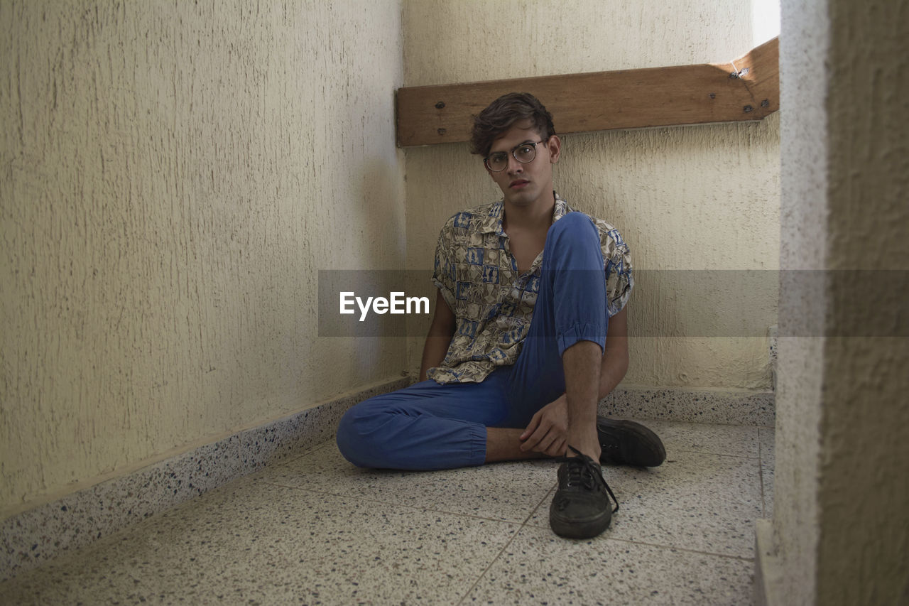 Full length portrait of young man sitting on floor against wall