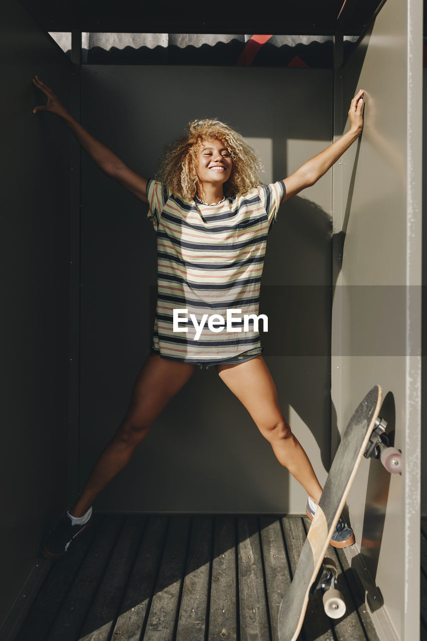 Cheerful female skateboarder with arms and legs outstretched amidst metallic walls on sunny day