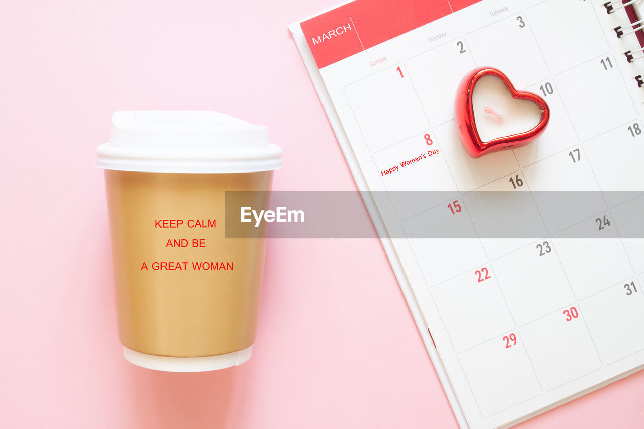 Directly above shot of disposable cup by calendar with heart shape against pink background