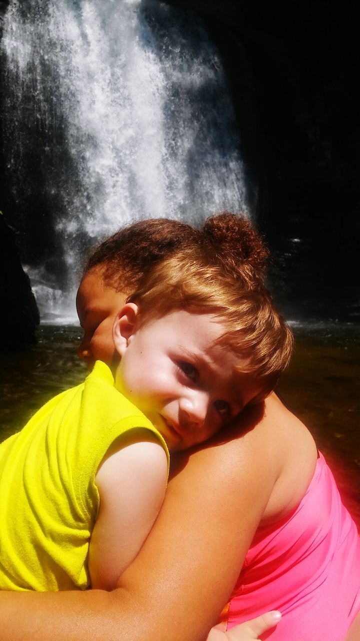 Side view of girl with brother against waterfall on sunny day