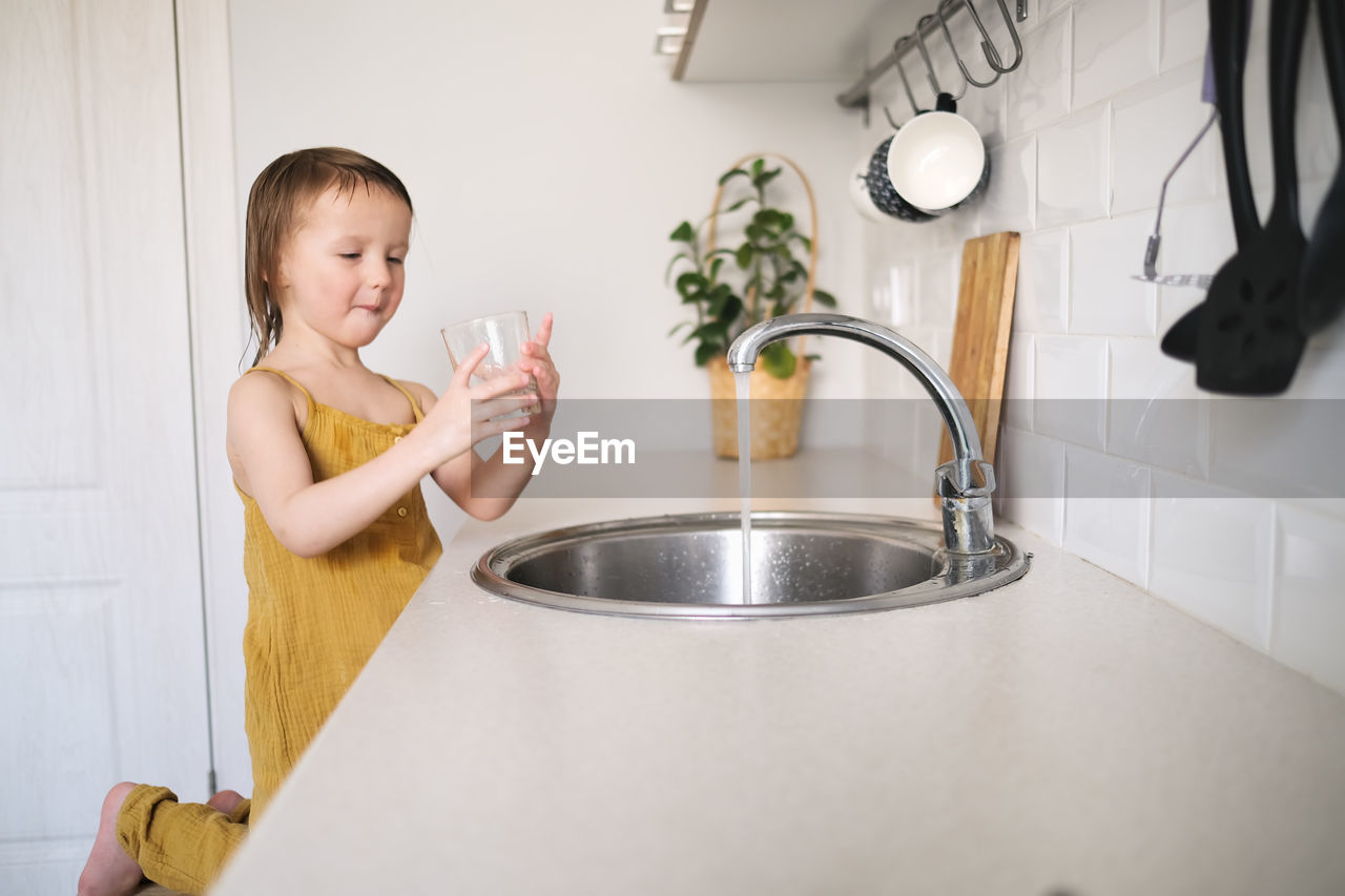 European child of 4 years old washes glass in sink on his own, bright kitchen in real interior, 