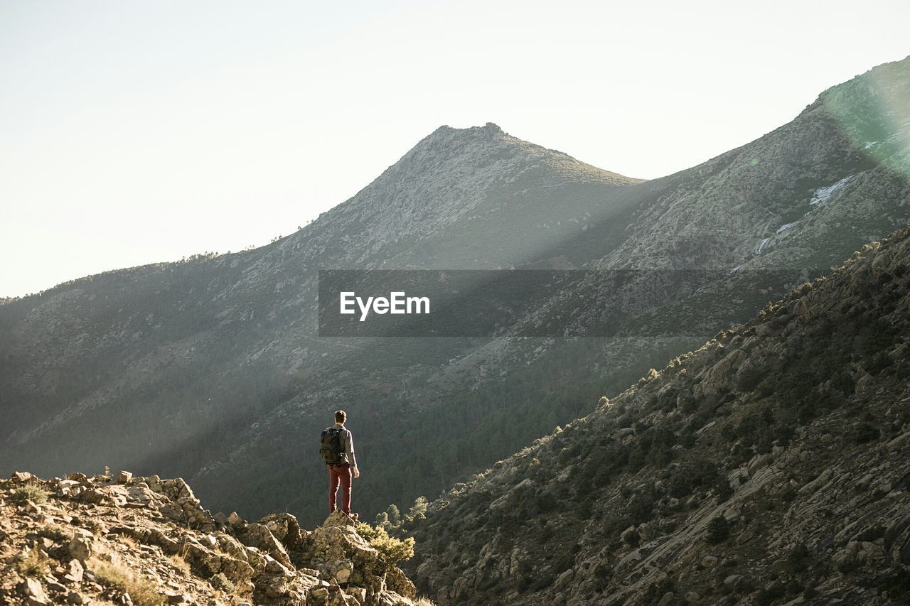 Rear view of man standing on cliff by mountains against clear sky