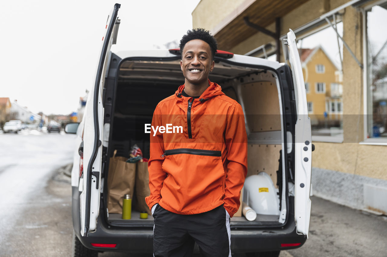 Portrait of smiling essential service man standing with hands in pockets against delivery van