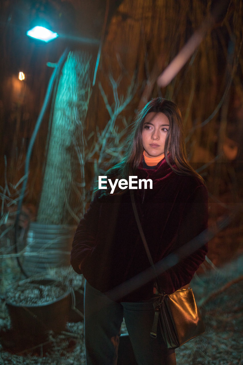 YOUNG WOMAN STANDING BY ILLUMINATED LIGHT AT NIGHT