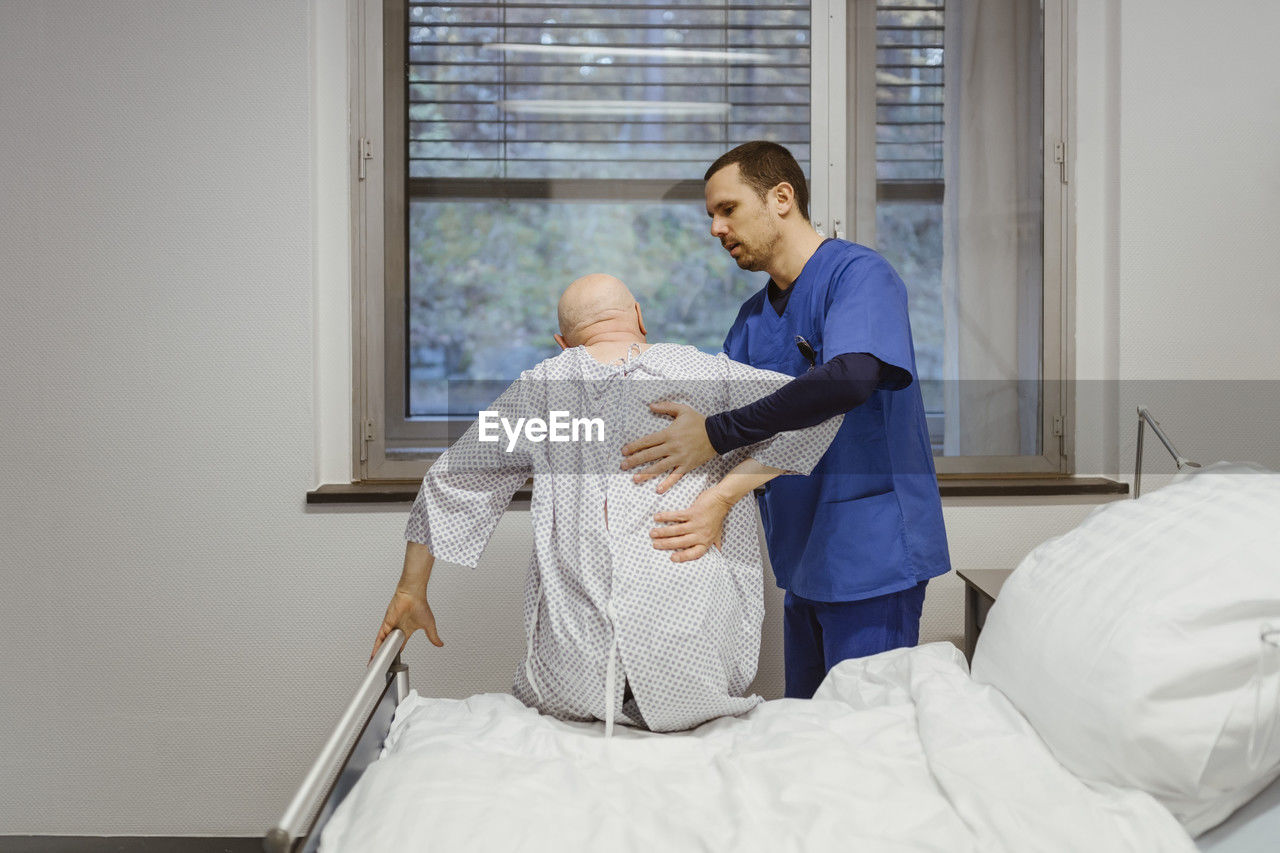 Male nurse helping senior patient sitting on bed at hospital