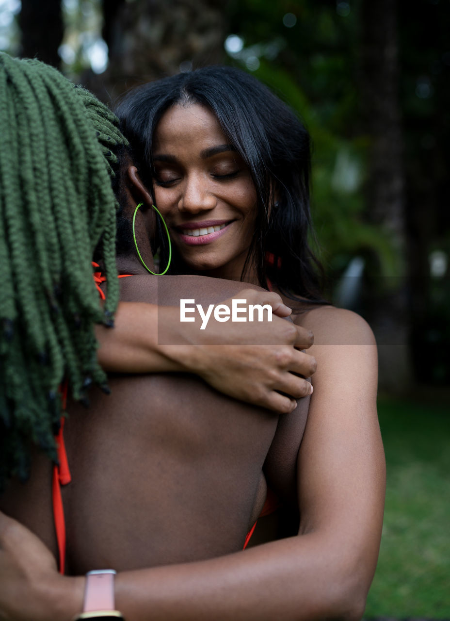 Content african american female friends in bikinis embracing while standing on lawn in backyard on summer day in tropical country