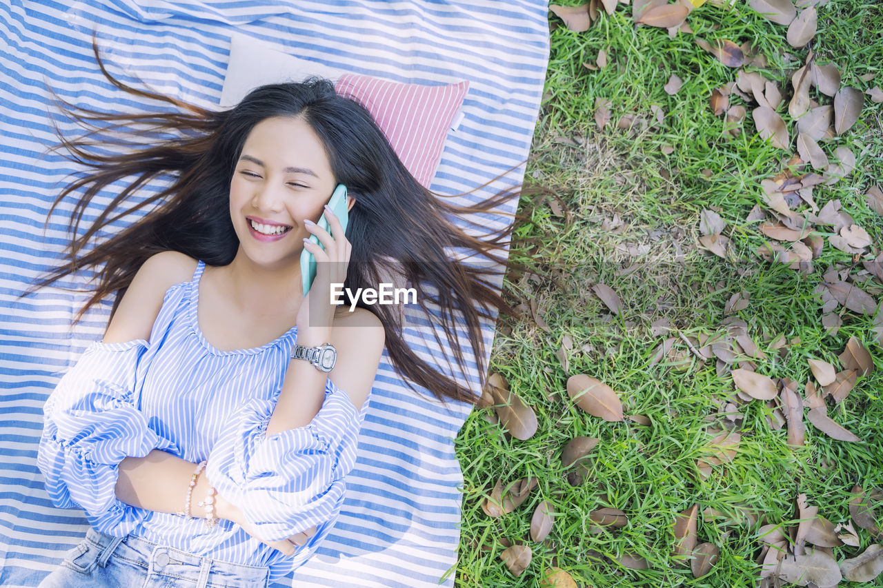 High angle view of smiling young woman talking over mobile phone while lying over blanket on grass