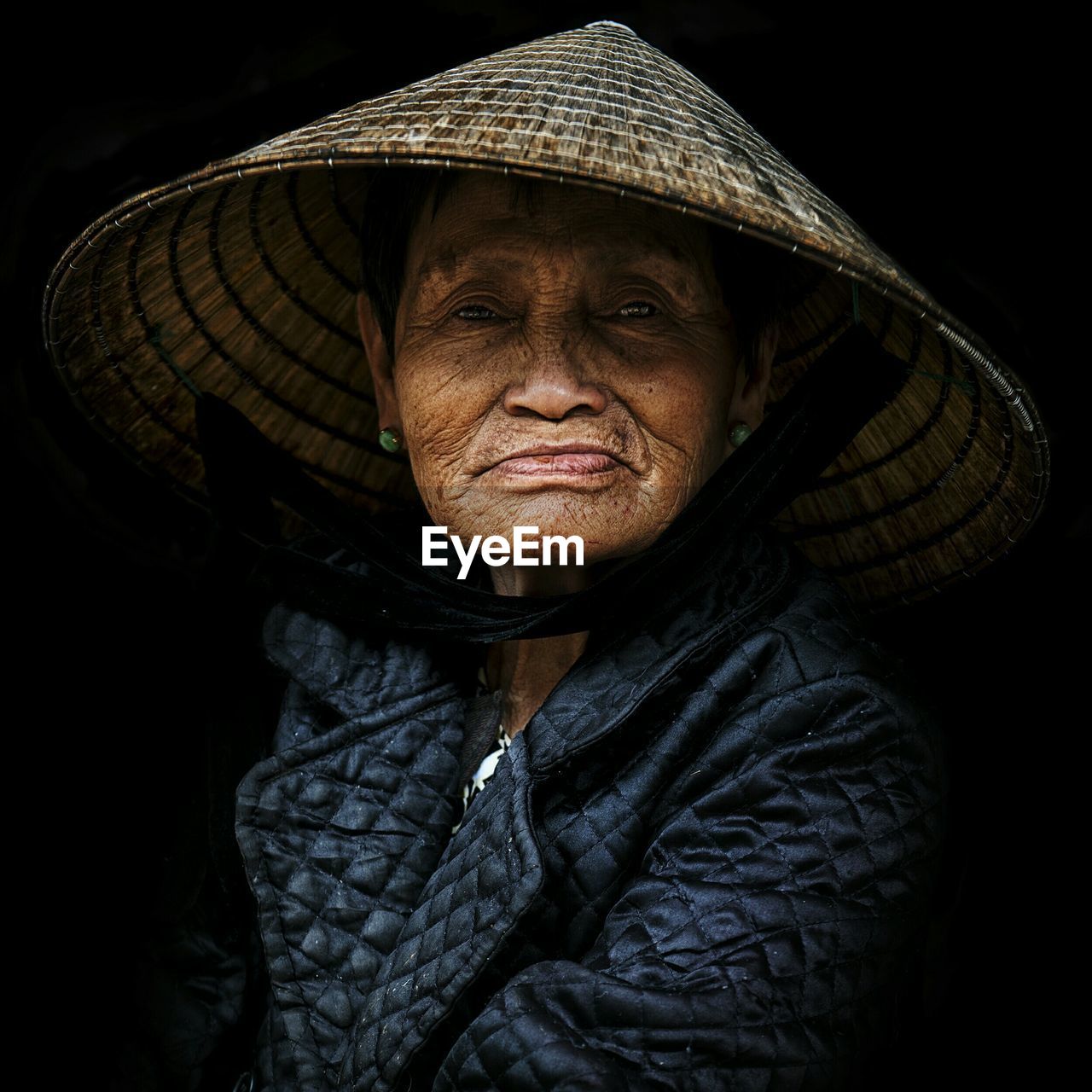 Portrait of senior woman wearing asian style conical hat against black background