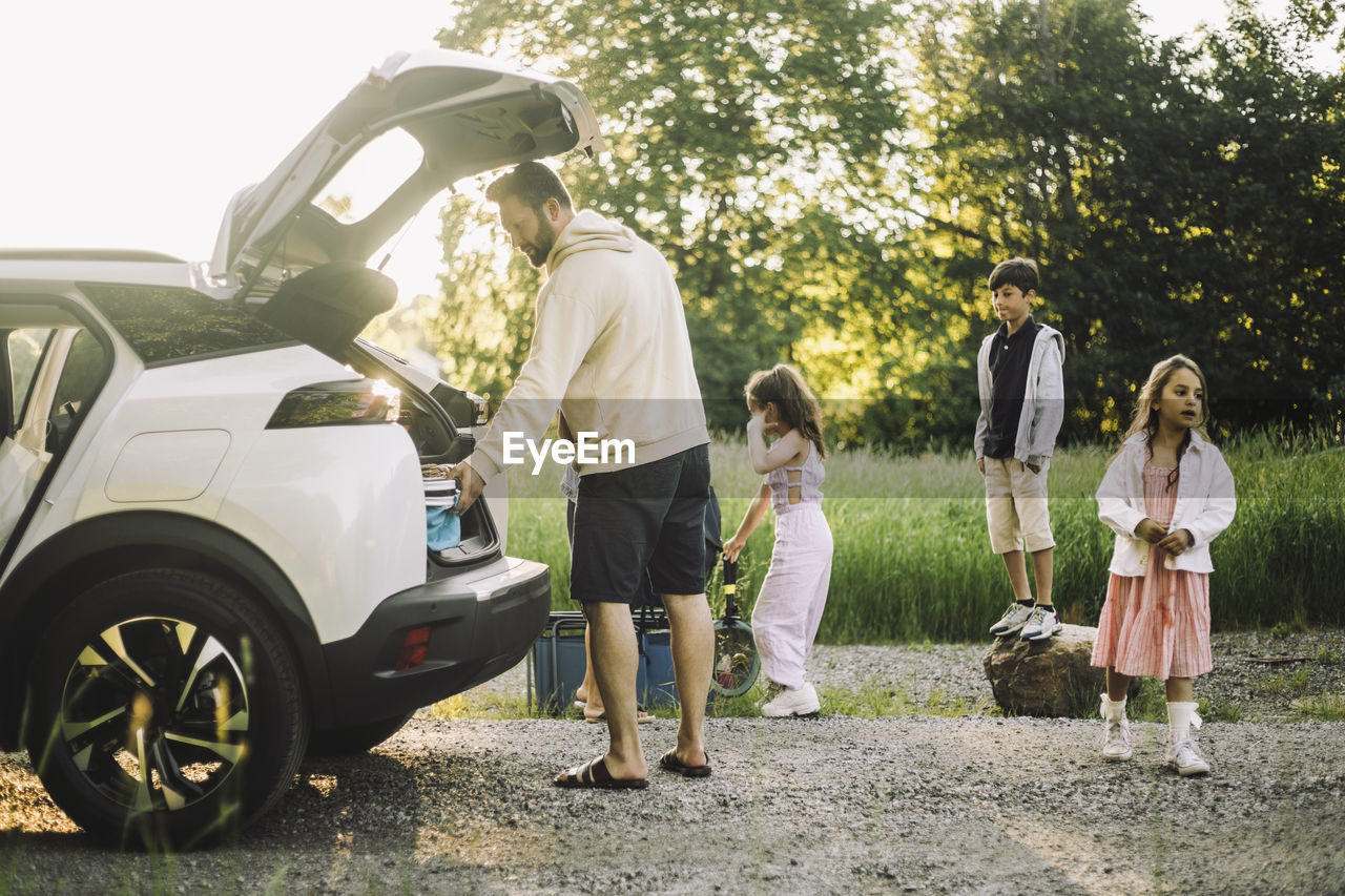 Father unloading stuffs from electric car trunk on dirt road