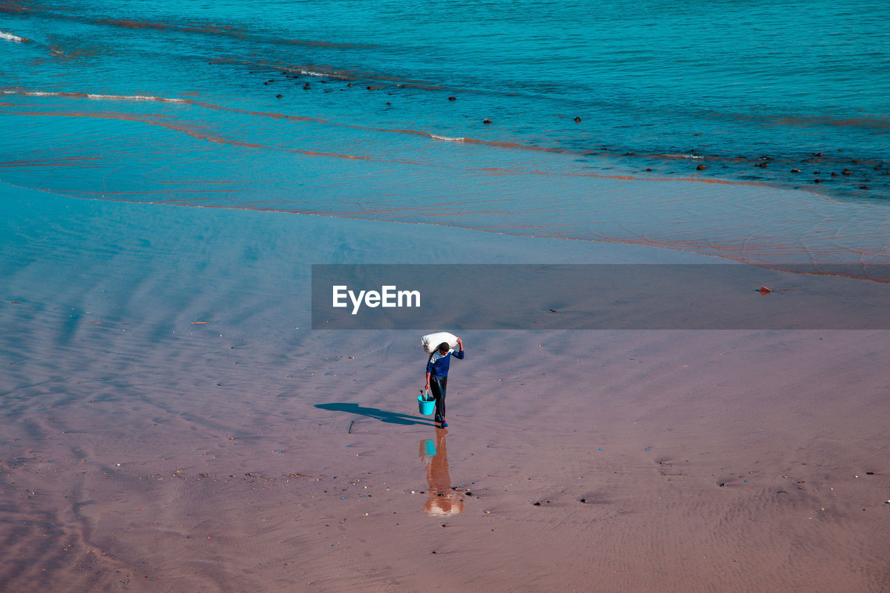 High angle view of man carrying sack on shoulders while walking on shore of beach